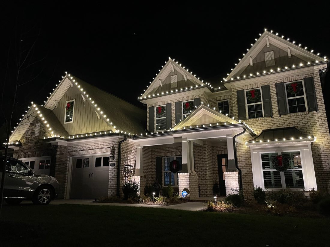 Craftsman-style home at night with C9 bulbs tracing four roof peaks and wreaths with red bows in every window.