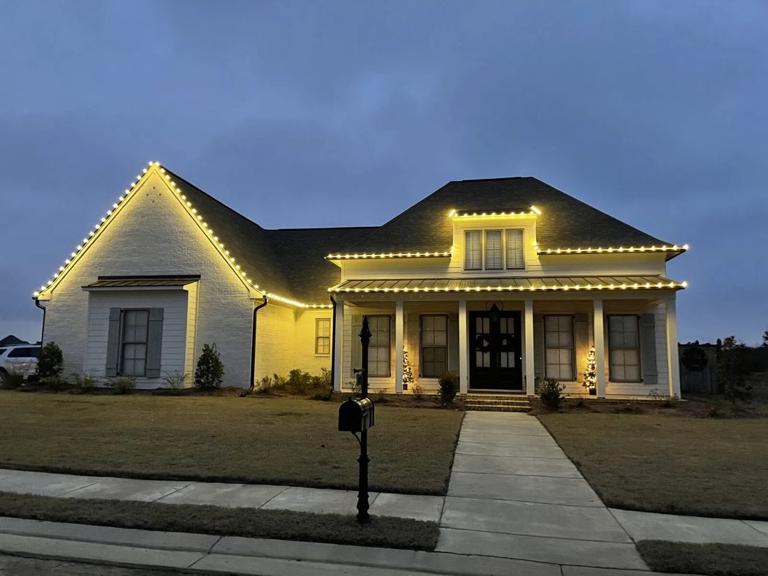 White farmhouse-style home at dusk with an evenly spaced warm white roofline and lit entry trees.