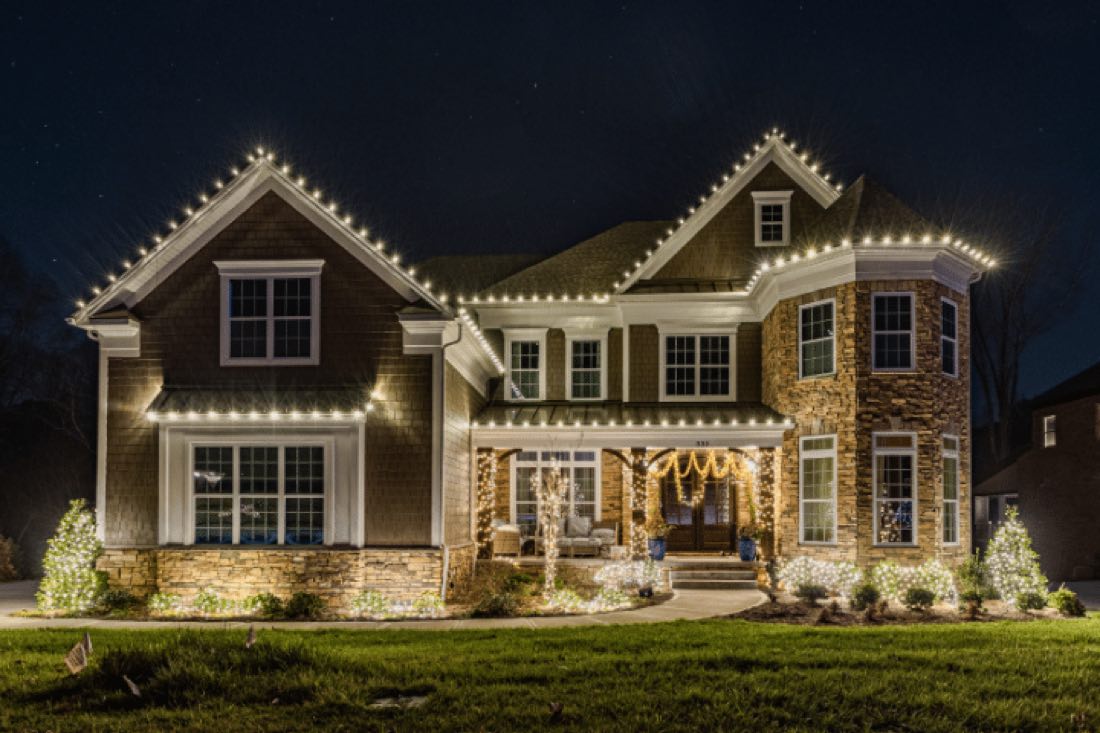 A two-story shingle-and-stone Connecticut home outlined in warm white roofline lights, with wrapped porch columns and lit shrubs.